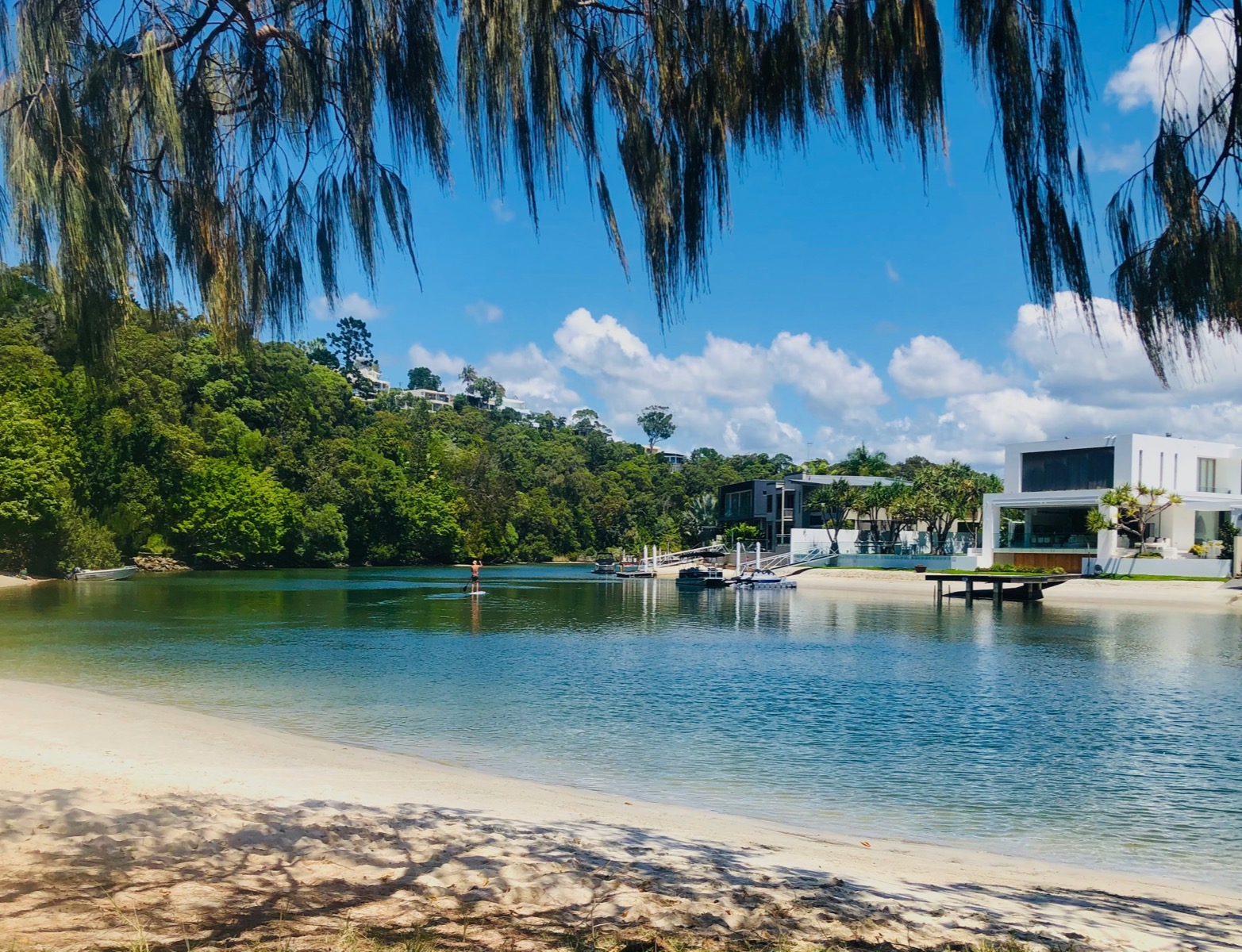 Paddleboarder crossing calm water between a sandy foreshore and waterfront homes
